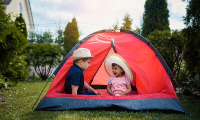Two happy little kids boy and girl brother and sister are playing in a red camping tent in the home yard on the grass.