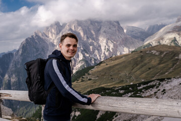 Naklejka premium Dolomites Alps. Italy. Smiling, positive brunet man with backpack lean on the wooden fence on background of green meadow, high mountain & blue sky in the clouds in sunny summer day