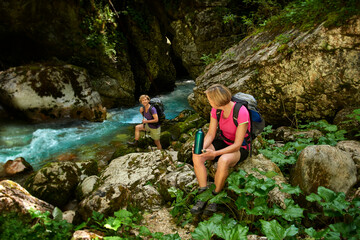 Two middle-age women are talking and resting by a alpine river taking a safe distance. Slovenia, Alps.