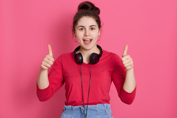 Happy excited female with dark hair and knot showing thumbs up and yelling something happily, posing in red shirt against pink background, lady having headphones around neck.