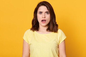 Astonished woman with pleasant appearance and dark beautiful hair, looking directly at camera, keeping mouth widely opened, expressive shock, looks surprised, wearing yellow t shirt.