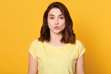 Fototapeta premium Portrait of lovely brunette female rounds lips as going to get kiss from boyfriend, looks at camera with pleased expression, isolated over yellow background.