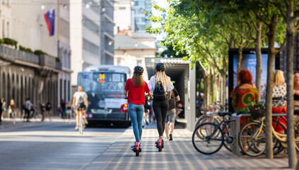 Rear view of trendy fashinable teenager girls riding public rental electric scooters in urban city...