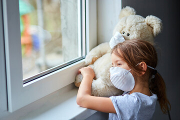 Little girl holding and hugging teddy bear in mask near the window.