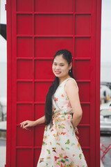 Close up portrait long hair Asian woman in a sweet beige long dress standing next to a red telephone booth with nature background.