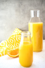 Obraz na płótnie Canvas A glass bottle and a decanter with freshly squeezed orange juice stand on a white surface against a gray concrete wall. Near lie one slice of orange and a yellow towel with a white pattern. Vertically