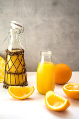 A delicious fresh orange juice in glass bottles stands on a white table against a gray concrete wall. One ethnic-style bottle is decorated with rattan. Near by lie the juicy slices of sweet orange.