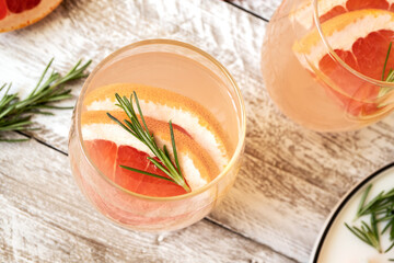 A refreshing drink with grapefruit juice in a glass stands on a light wooden table. In a glass slices of grapefruit and sprigs of rosemary. Summer fruit smoothies. The concept of soft drinks. 