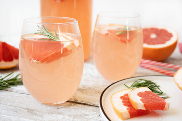 Two glasses of infused water with slices of grapefruit and rosemary stand on a light wooden table. Nearby is a white bowl with chopped grapefruit. The concept of summer refreshing drinks. Close-up