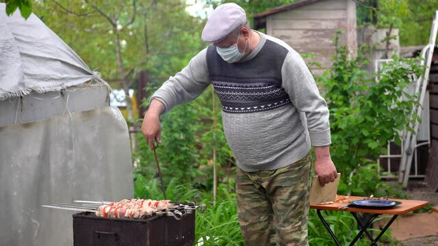 Mature man in medical mask impales raw shashlik on skewer. Older male holds skewer with barbecue. Concept of picnic outdoor.