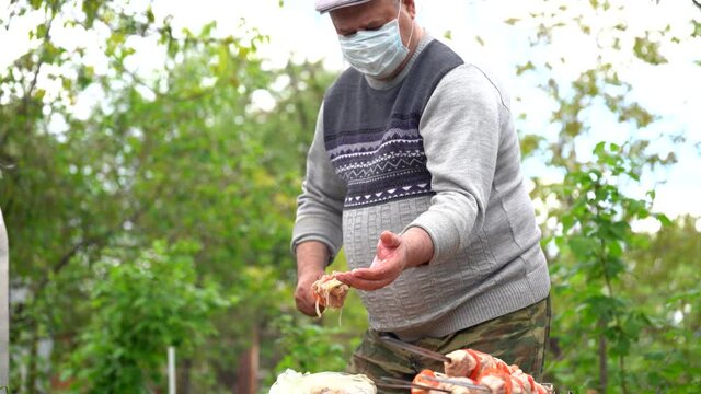 Mature man in medical mask impales raw shashlik on skewer. Older male holds skewer with barbecue. Concept of picnic outdoor.
