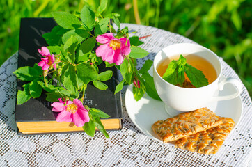 A Cup of green tea with a mint leaf, flowers, and a book on a table in the garden. Evening tea with cookies. Summer village concept. Vintage banner.