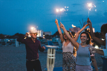 Happy friends having fun beach party outdoor with fireworks - Young people drinking champagne and dancing in summer vacation - Soft focus on left girl hand - Youth lifestyle and summer concept