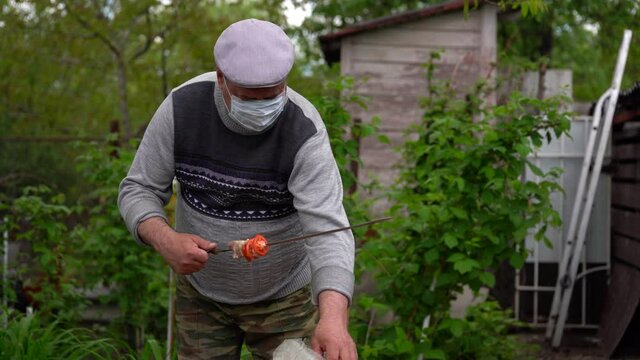 Mature man in medical mask impales raw shashlik on skewer. Older male holds skewer with barbecue. Concept of picnic outdoor.