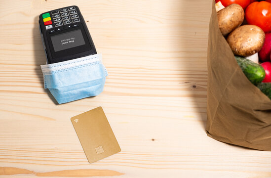 A POS with medical mask waiting for a contactless Nfc payment from gold card for a bag of groceries on a wooden background. The POS say: Stay Safe! Nfc pay. Covid pay. Safe pay .
