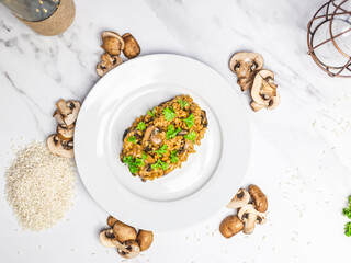 Mushroom risotto with Parmesan cheese and white Wine in a white serving plate on a white marble table with risotto Rice and raw mushroom on the side. Free space for your text/decoration.