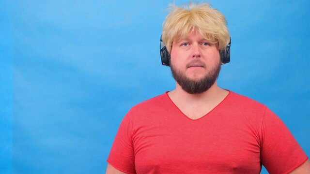 freaky fat man in a wig and a pink T-shirt in his headphones exhales cigarette smoke, smokes a vape against a blue background.