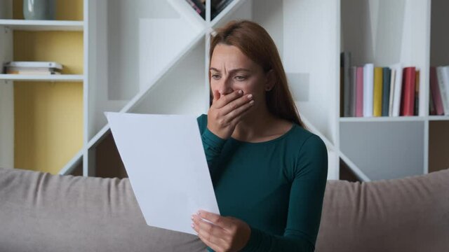 Sad woman sitting on couch at home reads received bad news holds documents paper letter feels desperate about financial problems, girl student worried college expulsion concept.