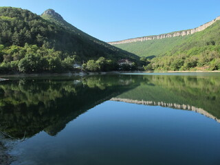 Fototapeta na wymiar The panorama of a mountain lake at the foot of a mountain plateau, the mirror surface of the water surface reflects high mountains and rocks, overgrown with dense green forest on a clear summer day. 