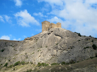 A picturesque panorama of the slope of a high rocky mountain in Sudak and the defensive towers of an ancient fortress against a bright blue sky on a sunny day.