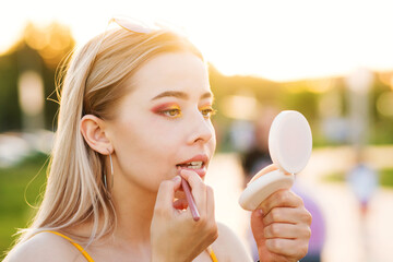 Stylish girl straightens bright makeup on the street. A young woman uses a lip liner and looks in the mirror of compact powder. Backstage. Close-up portrait in the sunset light. Hot summer evening