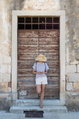 Fototapeta na wymiar Beautiful young female tourist woman standing in front of vinatage wooden door and textured stone wall at old Mediterranean town, smiling, holding, using smart phone to network on vacationes.