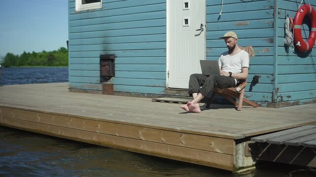 Bearded guy hipster is working with laptop and having nice day at lake pier on summer spbd. Young european man using computer for remote work and looking at screen while sitting by water. Portrait of