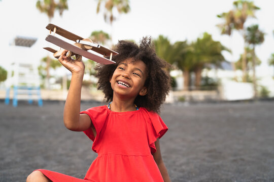 Afro child playing with wood toy airplane on the beach - Little kid having fun during summer holidays - Childhood and travel vacation concept
