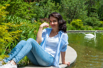 Fototapeta premium .Young brunette woman posing on a wooden pier near the lake. Girl dressed in jeans and t-shirt smiling. Swan lake, outdoor.