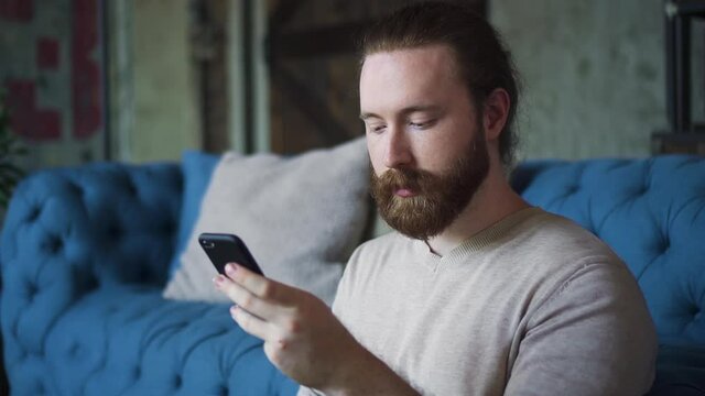 Portrait of young bearded guy is using smartphone and having good time at apartment room spbd. Handsome american man holding phone in hand and looking at screen, typing message. One male person uses