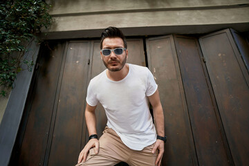 Stylish handsome young man with a bag near the metal gate, wearing white t-shirt with sunglasses 