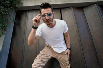 Stylish handsome young man with a bag near the metal gate, wearing white t-shirt with sunglasses 