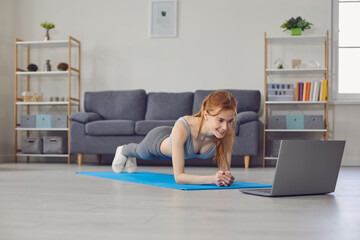 Online home sports. Pretty girl working out with laptop computer in living room. Woman making elbow plank exercise