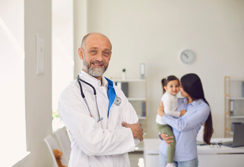 Fototapeta premium Smiling senior pediatric practitioner in medical office, with little girl and mother in background.