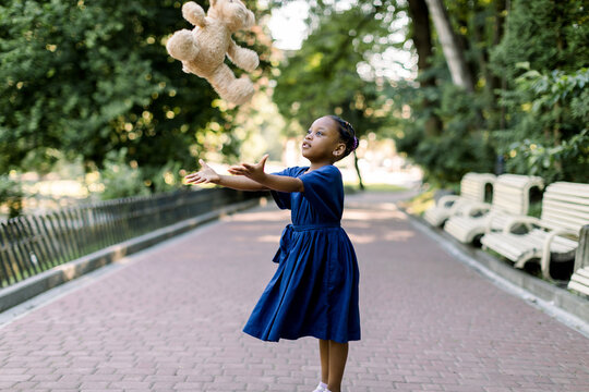 Portrait Of Happy Little African Girl Child In Green City Park Throwing Up Teddy Bear Toy, Flying In The Air. Smiling Dark Skinned Kid Girl In Blue Dress Playing In Summer Park.