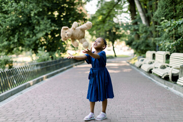 Cute and funny African kid girl wearing a colorful blue dress throws up a teddy bear in the park...