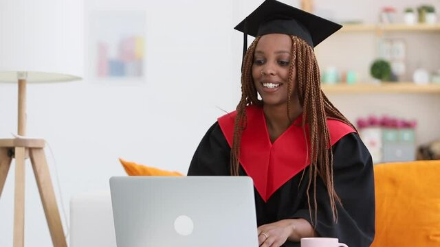 female student in cap and gown graduation costume Spbi and talking to grandparents online on a laptop. black afro woman sit in modern apartment and chat in online video call, greet and wave