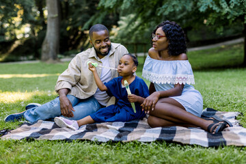 Fototapeta premium Family, parenthood and people concept. Happy smiling African family, father, mother and cute girl child in the park together on a sunny day, blowing soap bubbles