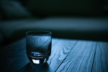 Glass on wooden table in empty dark room, sofa with pillows in background. Color grade in trendy...