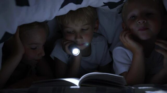 Three young children are reading a book with a flashlight under the covers at night.