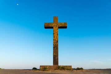 Big old Christian stone cross covered with bright yellow lichen in front of a clear blue sky at the beginning sunset. Concept of faith and religion giving hope. Low angle view, central perspective.
