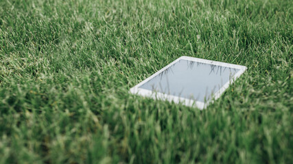 Close up top view of white tablet pc on the green grass in city park. Digital technology concept. Touchpad screen display of portable ebook with copy space. 5G network device. Mock up workspace.
