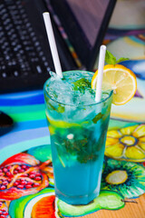 Blue cocktail with curacao and citruses and notebook background, working space. Alcoholic drink. Summer holidays.