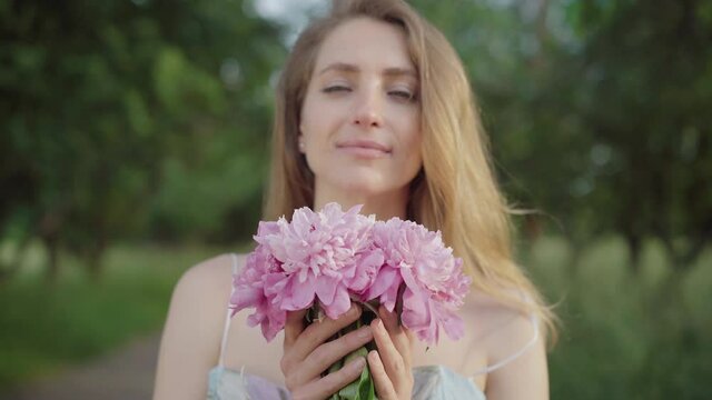 Beautiful young woman smelling bouquet of flowers in summer park. Portrait of charming Caucasian brunette lady enjoying weekend day outdoors. Beauty, leisure, lifestyle, joy.
