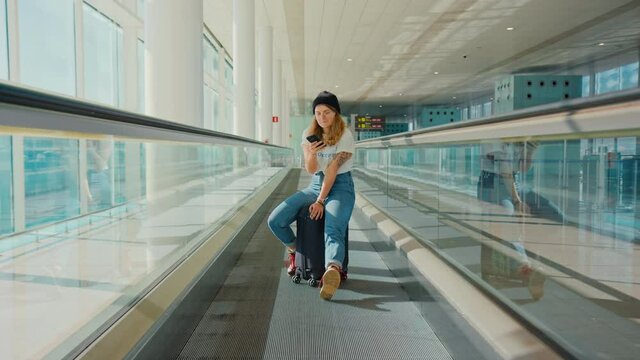 Trendy modern traveller, millennial young woman from generation z scrolls news feed on her smartphone, sit on suitcase while in airport. Cool teenager in empty airport terminal wait for flight