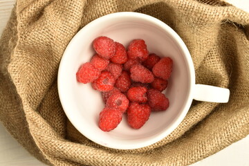 Juicy organic red raspberries in a white cup, close-up, on a jute fabric.