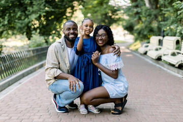 Summer family portrait in nature outdoors. Beautiful young African American family with cute little daughter, posing in park alley, hugging each other and looking at the camera