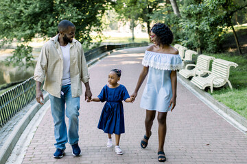 Happy African family, father, mother and cute little child girl, holding hands while walking together in beautiful city park alley with trees and sunset on the background, having time together.