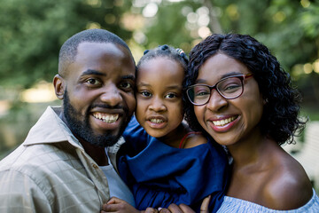 Close up portrait of young African American family with little cute daughter in the summer park. Smiling family looking at camera, enjoying walk in nature