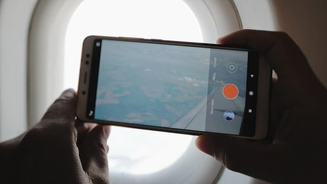 A man takes pictures of a wing of an airplane and an airplane engine from a porthole window using a camera smartphones white.